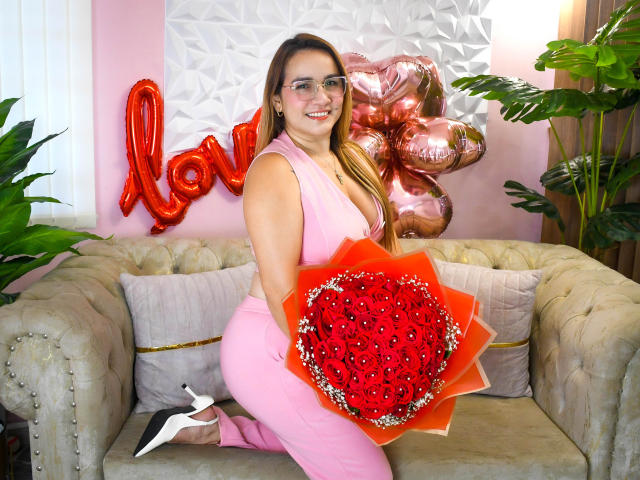 ManuelaMature, with glasses and a pink dress, kneeling on a couch, holding a red heart bouquet, smiling, pink high heels, and pink balloons behind her.
