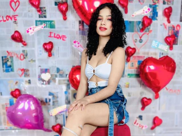 BellainLove sits on a red stool, wearing a white bra with a bow and denim shorts. She has curly black hair and red lipstick. Her skin is light brown. Red and pink heart-shaped balloons are scattered around her, with "I LOVE" written in pink on the wall. The wall is covered in newspaper and small photos. Her left knee is bandaged. The composition is bright and playful, with a focus on romantic and casual elements. The image has a modern, pop-art feel.