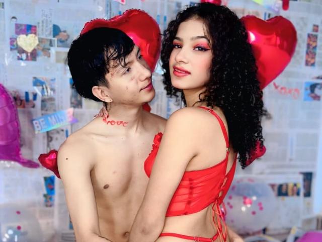 A photo of BellainLove and another person. BellainLove has curly black hair, red lipstick, and wears a red bra with heart-shaped accents. The other person has short black hair, a tattoo on their neck, and is shirtless. Both are looking at each other, with red heart-shaped balloons in the background. The red bra contrasts with BellainLove's pale skin. The image uses vibrant colors and romantic themes, with a focus on the subjects' expressions and red accents.