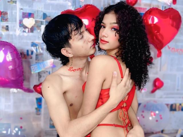 BellainLove with curly black hair, red lingerie, and pink lipstick, being hugged by a shirtless guy with black hair. Red heart balloons and pink balloon in the background.