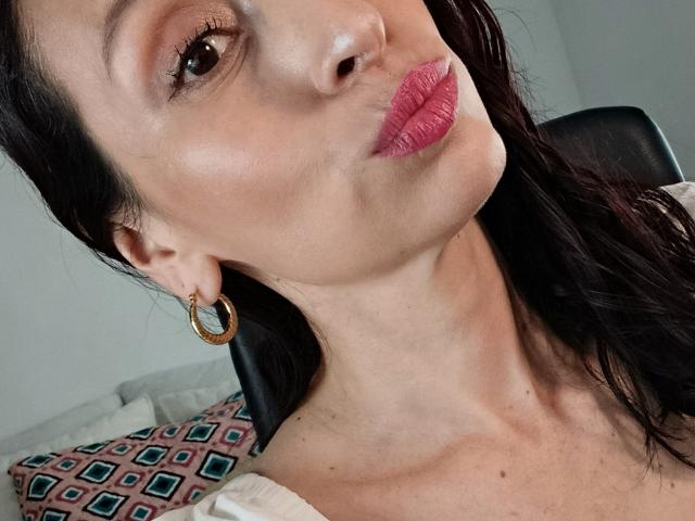 Close-up of AliceRossi's face, showing a slight pout. She has fair skin, dark brown hair, and is wearing pink lipstick. Her right ear has a gold hoop earring. She's looking to the left with her head tilted. She has subtle makeup with light eyeshadow. The background is blurred, featuring a patterned pillow and a black chair. The image focuses on AliceRossi's facial features and jewelry.