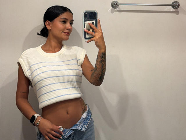 ThaliaAres with dark hair in a ponytail, wearing a white striped crop top, blue jeans, and a wrist bracelet, taking a mirror selfie, showing her tattooed arm and flat ass.
