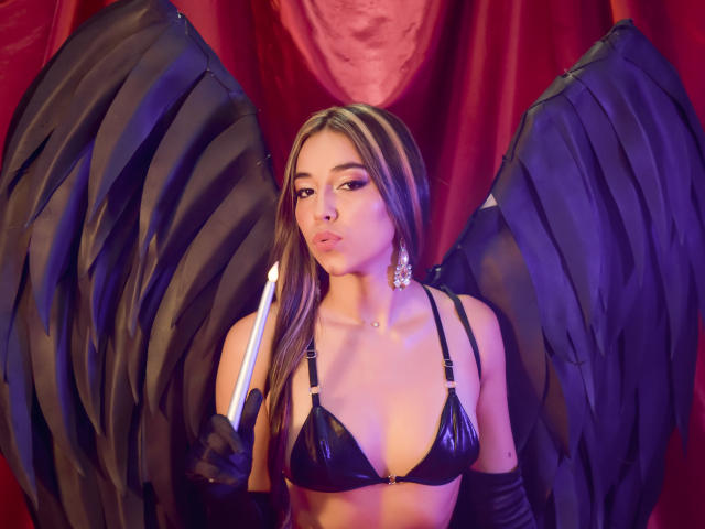 MissVallen with black wings, wearing a shiny black bra, holding a sword, and a diamond earring, has pouty lips and long brown hair. Her tits are visible.