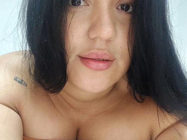 LauraRichis with black hair, pink lips, and a small tattoo on her right shoulder, looking at the camera, showing her bare boobs. Her skin is light, and she's topless.