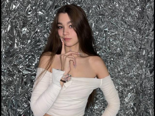 MerrieWirebaugh has long brown hair, light skin, and a slight smile. She's wearing a white off-shoulder, long-sleeve top that fits snugly. Her right hand is raised with fingers touching her chin. The background is filled with crinkled, silver foil, creating a chaotic texture. She has a delicate, natural look with subtle makeup. Her nails are painted a light color. The composition is centered on MerrieWirebaugh, who stands slightly to the right. The overall style is modern and edgy.