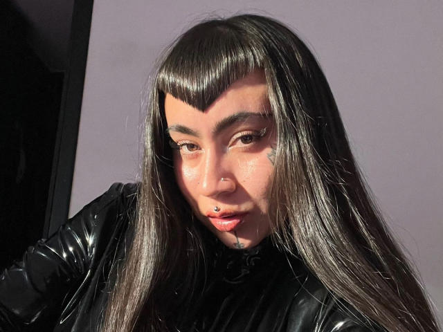 Close-up of Jesskhy with long, straight black hair and blunt bangs. Jesskhy has a light brown skin tone, brown eyes, and wears winged eyeliner. They have a small nose ring on the left side of their nose and a visible lip piercing. Jesskhy is wearing a shiny black leather jacket. The light is bright and casts shadows on their face. The image is sharp with a realistic texture to the hair and leather. The overall composition is centered on Jesskhy's face and upper torso.