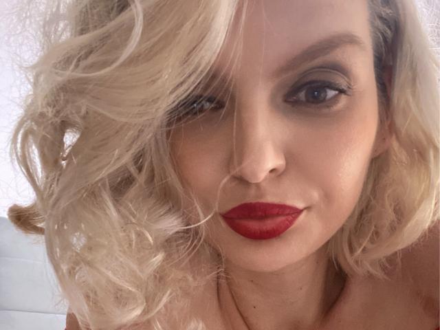 DouceAmelia has blonde curly hair, red lipstick, and a slight smile. Her blue eyes are slightly squinted, and she's wearing a necklace. Her tits are not visible.
