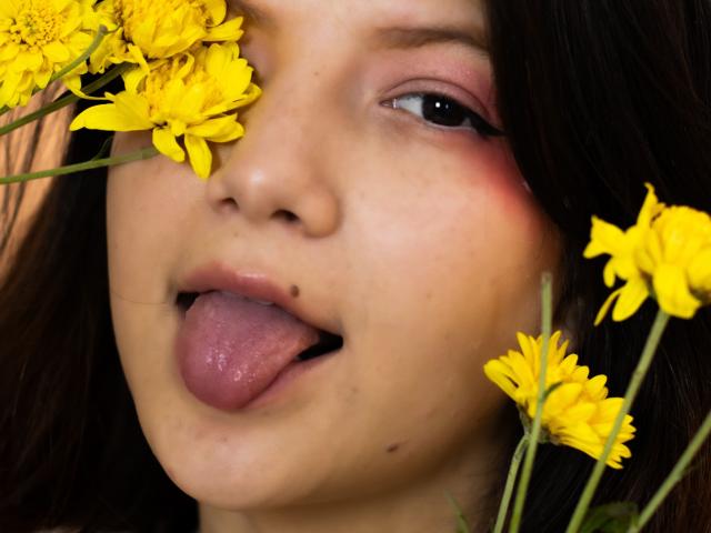 MiaBellee's face with yellow daisies, tongue out, dark hair, pinkish eyes, and a small beauty mark on her left cheek, showing a playful and cute expression.