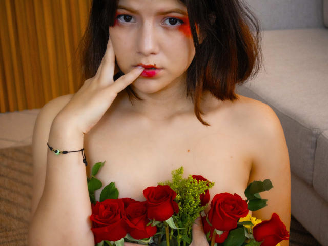 MiaBellee with red rose "boobs" covered, hand near mouth, finger touching lips, dark brown hair, red eyeshadow, pale skin, gray couch, black bracelet, green stem, yellow flower.