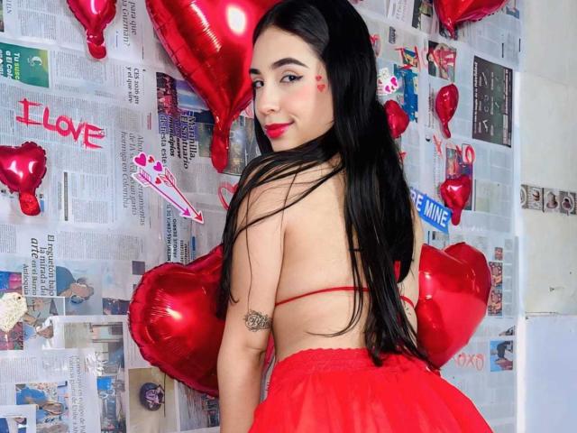 ArdienteCouple with long black hair, red lipstick, and red heart tattoo, wearing a red strapless top and skirt, looking back with a smile, surrounded by red heart balloons.