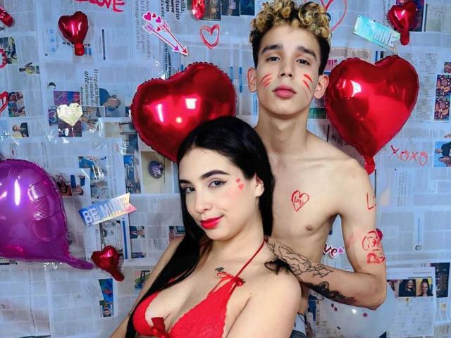 ArdienteCouple, a shirtless dude with curly blonde hair and red face paint, stands behind ArdienteCouple, a Latina chick with red lingerie and black hair, holding red hearts.