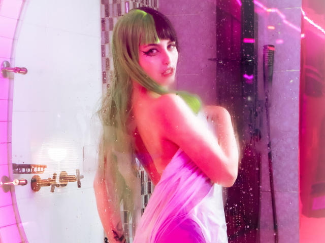AngelBlakee has long green and black-dyed hair, standing in a shower with pink neon light reflecting on the wet glass. She's facing slightly to the right, covering her small breasts with her arm. Her makeup is bold, with thick black eyeliner. She's wrapped in a white towel that's partially falling off her shoulders. The shower has white tiles, brass faucets, and a visible showerhead. The image has a slightly blurred, almost digital effect, with visible water spots on the glass.
