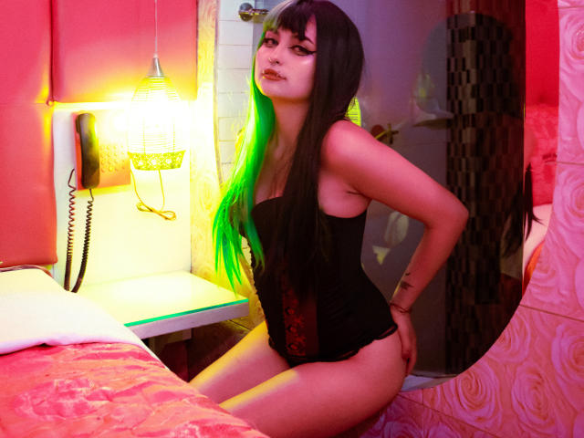 AngelBlakee has long black hair with bright green highlights, wearing a black strapless corset, sitting on a bed with red and pink patterned sheets. She has a tattoo on her right wrist and is looking to the side with a slightly open mouth. A yellow lamp with a textured shade and a wall-mounted phone are in the background. The room has a mix of red, pink, and white colors. The light from the lamp casts a green glow on her hair. The composition focuses on her and the bed.