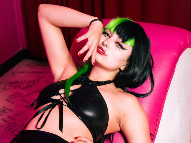 AngelBlakee with green and black hair, black leather bra, nose ring, and red nails, lying on pink chair, hand on forehead, smiling, showing her tits.