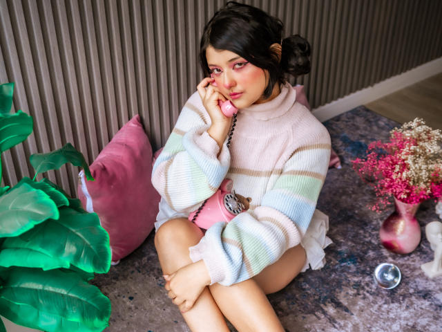 MisakiWong sitting on floor, wearing pastel striped sweater, holding pink phone to ear, pink blush, green plant, pink pillow, pink flower vase, and silver ring on carpet.
