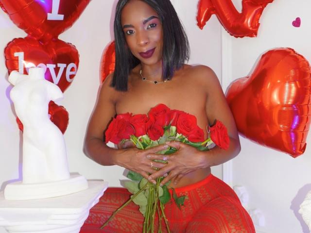 JadaMillss with dark brown hair, red lipstick, and black necklace, wearing red lace pants, holding red roses, and sitting in front of a white statue, surrounded by red heart balloons.