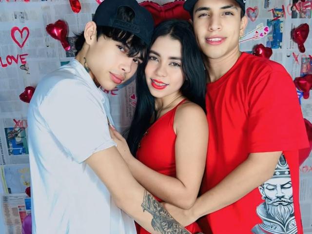 Three people, InocentLooks with dark hair, red dress, and tattoos, flanked by two guys in white and red shirts, holding each other, smiling. Red heart balloons in background.