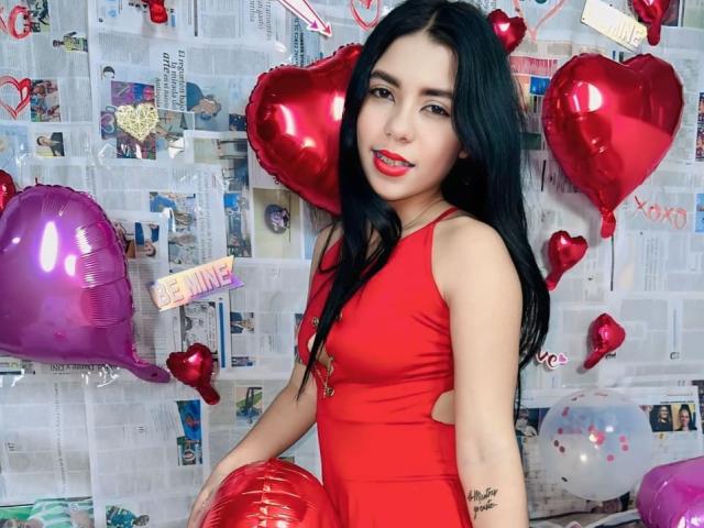 InocentLooks with black hair, red lipstick, and red dress, holding red heart balloon, standing with purple heart balloon, showing tattoo on right arm, smiling.