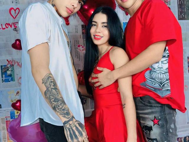 InocentLooks in a red dress, standing between two guys. One guy in a white shirt, tattooed arm, touching her tits. Other guy in red shirt, hand on her back.