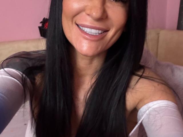 AndraD with long black hair, smiling, showing white teeth, wearing a white off-shoulder top, pinkish skin, and visible cleavage. Boobs are partially visible. AndraD has a sexy look.