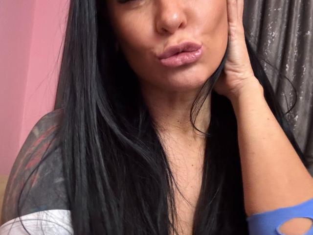 AndraD has long black hair, pouty pink lips, and a tan skin tone, with her hand on her cheek, wearing a blue and gray top, showing a cut-out.