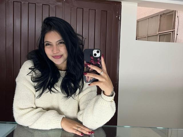 NyraVanel mit schwarzen Haaren, einem weißen Pullover und roten Nägeln, lächelt in ein Spiegel-Selfie, hält ein Handy mit einer schwarzen Hülle, im Hintergrund eine braune Tür..