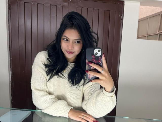 NyraVanel, con lunghi capelli neri, indossa un maglione crema, tiene in mano un telefono con una custodia a fiori, sta scattando un selfie allo specchio, sorride leggermente, ha le unghie rosse, ed è in piedi davanti a una porta..