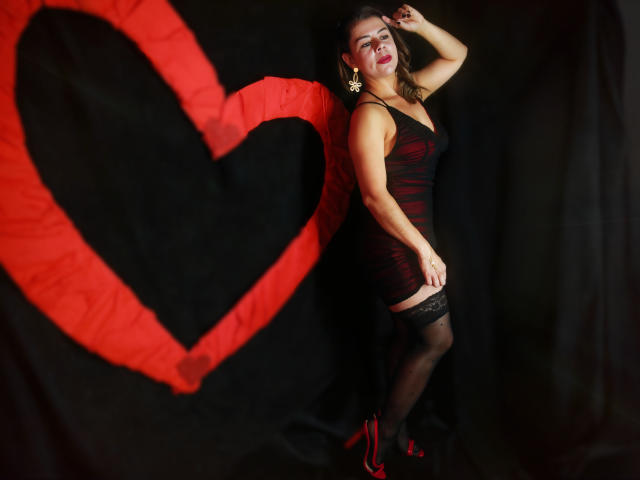EmiliaMilf in a black, red-striped dress, black thigh-high stockings, and red heels, posing with hand in hair, standing next to a large red heart on a black backdrop.