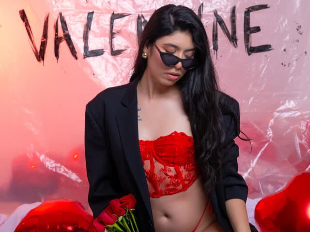 MiluSky, avec ses longs cheveux noirs, portant des lunettes de soleil noires, un soutien-gorge en dentelle rouge, un blazer noir et un string rouge, tenant des roses, et avec un tatouage sur sa poitrine. Ses seins sont visibles..