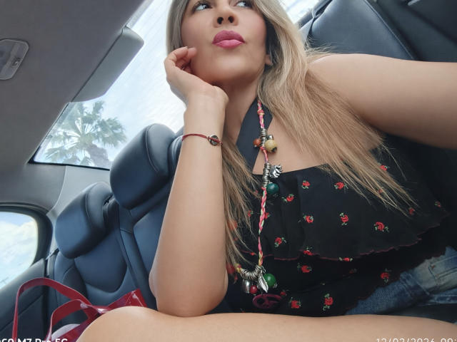 Maytena with long blonde hair, pink lipstick, and black floral dress, sitting in car, hand on cheek, red bracelet, necklace, and red bag, looking up with a cute expression.