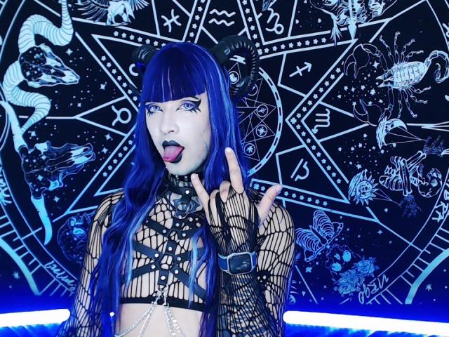 NaomyWoman with long blue hair, black fishnet top, and straps, sticking out tongue, wearing black horn headpiece, black choker, and fingerless gloves, against a celestial, magical pattern.