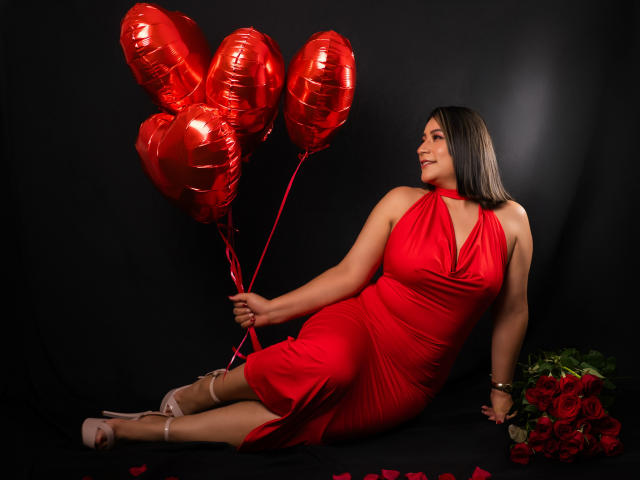 EmaFoxy indossa un abito rosso, tiene in mano palloncini a forma di cuore rossi e lucidi, è seduta tra le rose e indossa dei tacchi rosa, sorridendo di lato. Il suo sedere è coperto dall'abito..