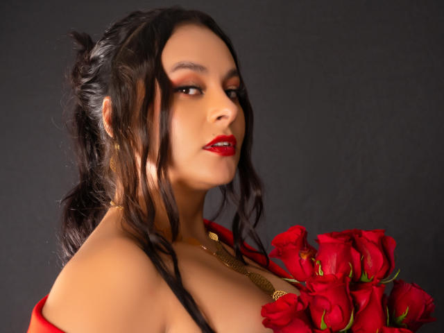 LunaSoler trägt roten Lippenstift, hat dunkles, zu einem unordentlichen Pferdeschwanz gebundenes Haar und hält rote Rosen. Sie trägt eine goldene Halskette, und ihre Brüste sind teilweise gegen einen dunklen Hintergrund sichtbar..