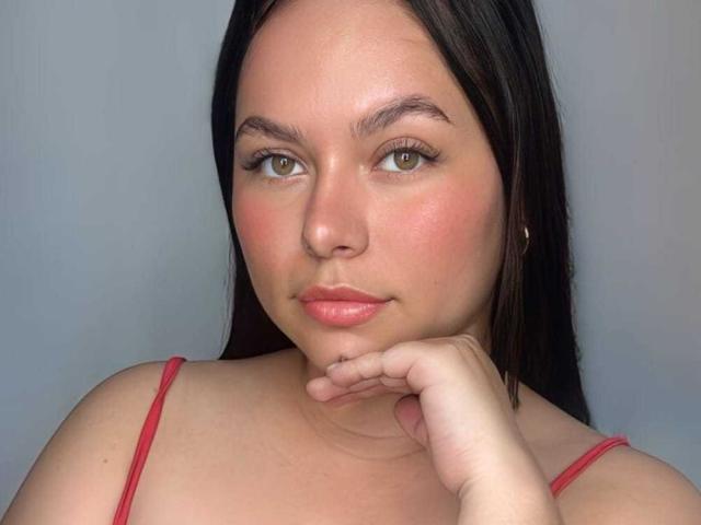 SamyCastiel69 has pale skin, green eyes, and pink lips. She's resting her chin on her hand, wearing a red spaghetti strap top, with long black hair.