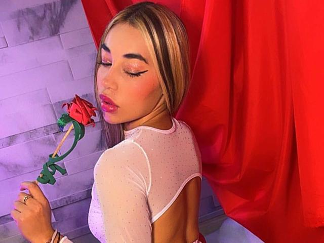 MissVallen with blonde hair, pink eyeliner, and pink lipstick, holding a red rose, wearing a sheer white top, back to the camera, red curtain in the background.