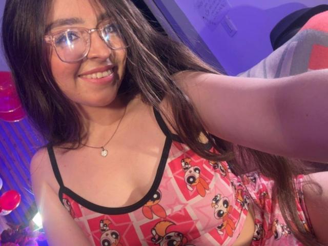 EvaMcAllister with long brown hair, glasses, smiling, wearing pink Mickey Mouse crop top and matching shorts, showing her tits, in a purple-lit room, taking a selfie.