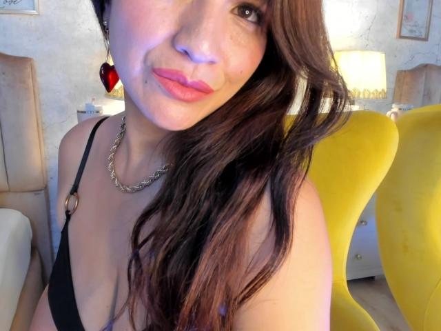 CameronViviane with wavy brown hair, pink lips, black strap dress, silver chain necklace, red heart earrings, yellow chair, and light skin, smiling in a well-lit room.