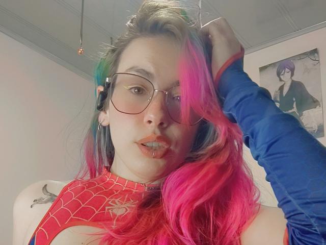 NatashaLau with pink and green hair, glasses, red Spider-Man top, blue arm sleeve, and tongue piercing, holding hair, looking at camera.
