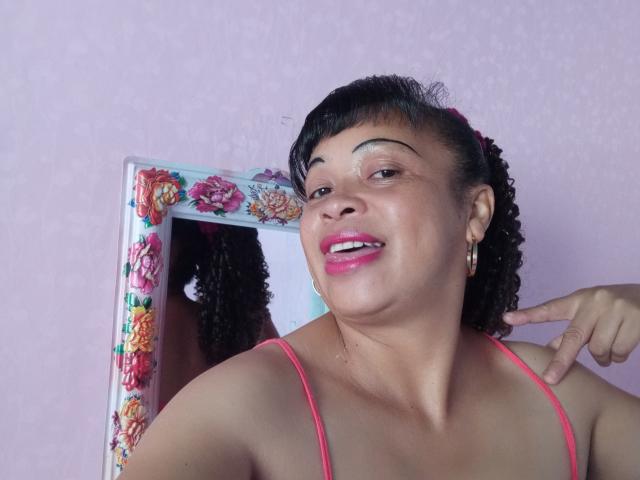 Rahel69 with curly black hair, pink lipstick, and pink spaghetti straps, pointing to her cheek with a smile, reflected in a floral-framed mirror.
