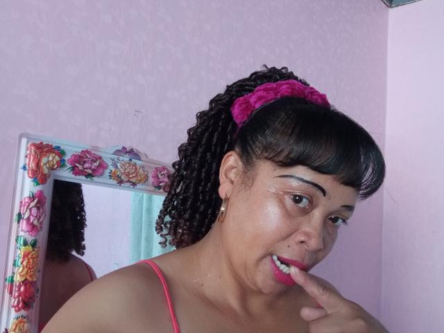 Rahel69 has a medium-brown complexion, curly black hair in a ponytail with a pink scrunchie, and wears a pink tank top. She's licking her pink-lipsticked finger with a slight smile. Her eyebrows are dark and defined, and she has small earrings. A floral-framed mirror is to her left. The pink wall has a subtle pattern. The photo is focused on her upper body and face, capturing a casual, self-reflective moment.