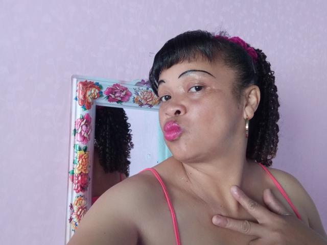 Rahel69, dark curly hair, pink scrunchie, pink spaghetti straps, pouty pink lips, hand on chest, mirror with floral frame, pink wall, earrings, light brown skin, looking at camera.
