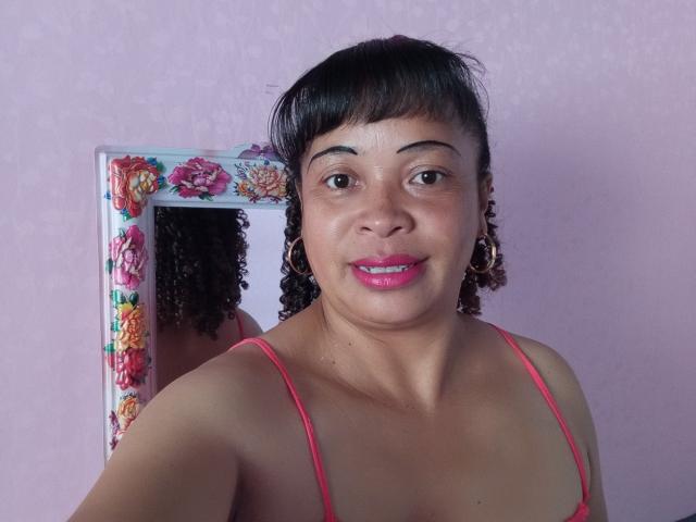 Rahel69 has medium brown skin, black curly hair, pink lipstick, and wears pink spaghetti straps. She's in front of a floral mirror, smiling at the camera.