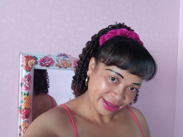 Rahel69 with curly black hair, pink scrunchie, pink lipstick, and pink spaghetti straps, smiling at the camera, reflected in a floral-framed mirror. Her tits are partially visible.
