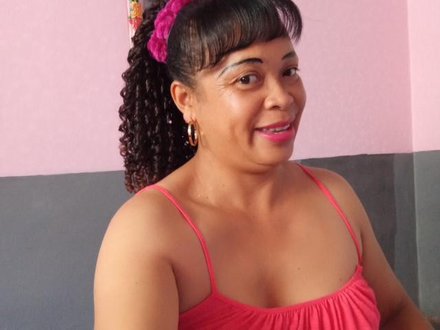 Rahel69 with curly brown hair in a pink scrunchie, wearing a pink spaghetti-strap top, smiling, showing small tits, brown skin, pink lipstick, gold hoop earrings, and a cute face.