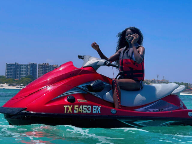 A red jet ski with "TX 58453 BK" on the side. KharlaQueen, with dark curly hair and wearing a red life jacket, sits on the jet ski. She's holding a phone to her ear with her right hand. Clear blue water and a distant city skyline in the background. Bright sunlight highlights the jet ski's glossy surface. KharlaQueen's life jacket contrasts with the red jet ski.