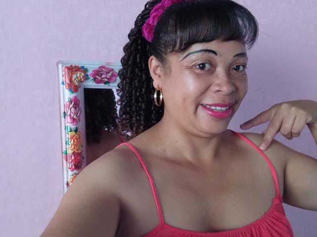Rahel69 with curly black hair in pink scrunchie, pink lipstick, hoop earrings, and pink tank top, pointing at chest, smiling. Reflective floral mirror behind her.