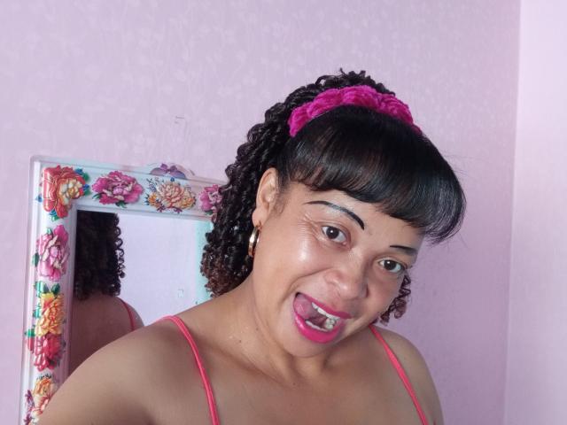 Rahel69 with curly black hair, pink scrunchie, and hoop earrings, sticking out her tongue, pink spaghetti strap top, reflected in a floral-framed mirror, pink walls.