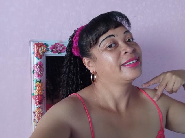 Rahel69 with medium brown skin, black curly hair in pink scrunchie, pink lipstick, gold hoop earrings, pointing at her chin, wearing pink spaghetti straps, smiling.