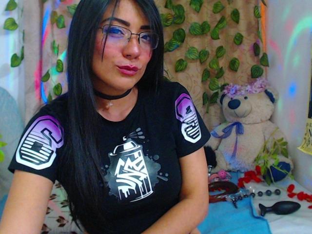 SophiQueen with long black hair, glasses, pink lipstick, black choker, black t-shirt with purple and white graphics, sitting with a cute white teddy bear and sex toys on a bed.