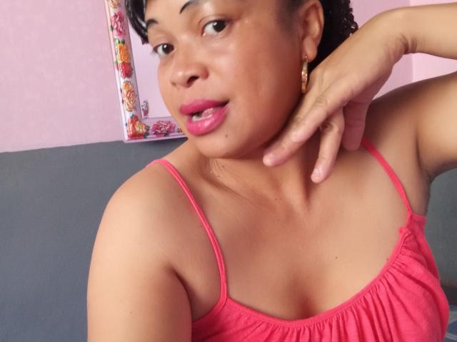 Rahel69 with dark skin, pink lipstick, and curly black hair, wearing a pink spaghetti-strap top, has her chin raised and hand touching her neck, looking slightly open-mouthed.